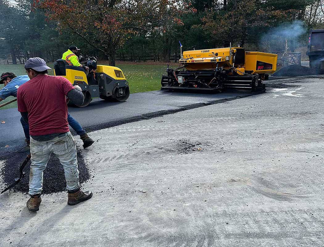 Workers pave a road; one spreads asphalt while another drives a roller. A yellow paving machine lays fresh asphalt, and trees with autumn leaves are visible in the background.