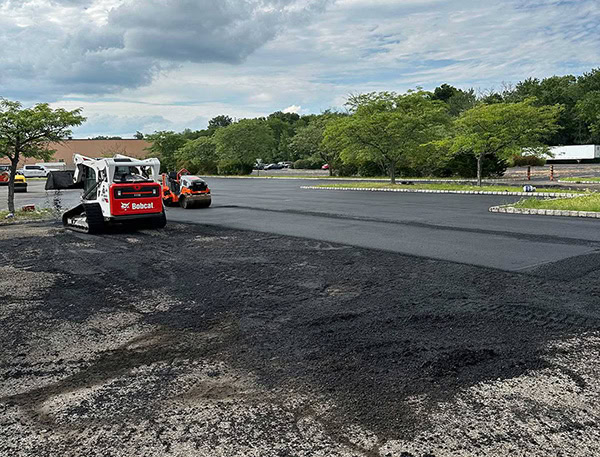 a&s-paving-during-the-job Construction workers use a Bobcat and a roller to pave and smooth fresh asphalt on a large parking lot surrounded by trees and parked cars under a partly cloudy sky.