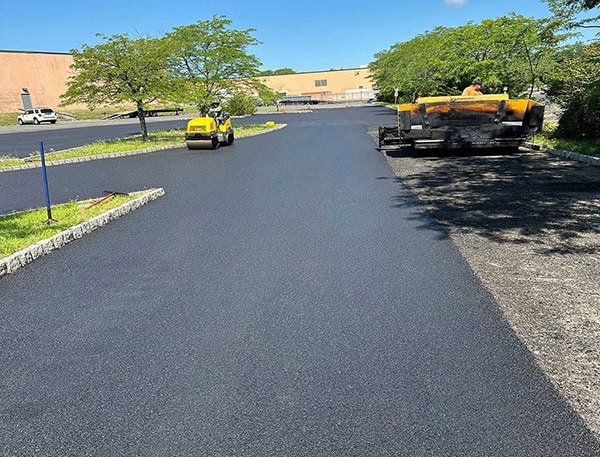 a&s-paving-on-the-job Freshly paved asphalt parking lot with construction vehicles and workers present. Trees line the edge, and a large building is visible in the background under a clear blue sky.