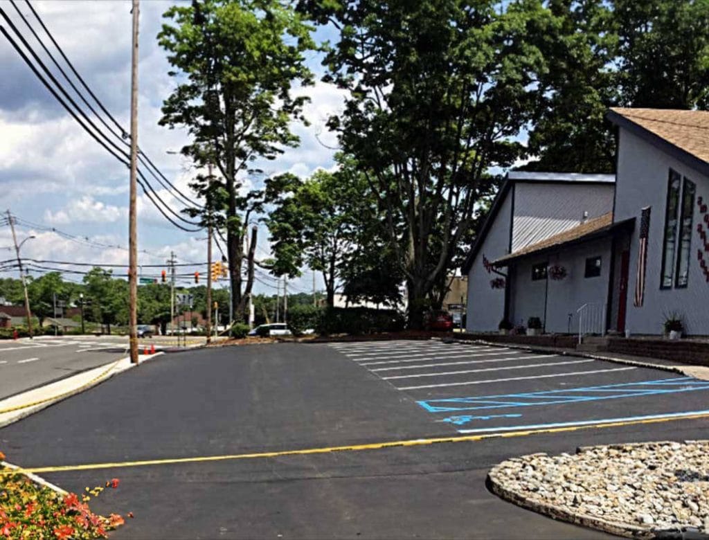 Freshly paved, empty parking lot beside a building with trees and a road in the background.