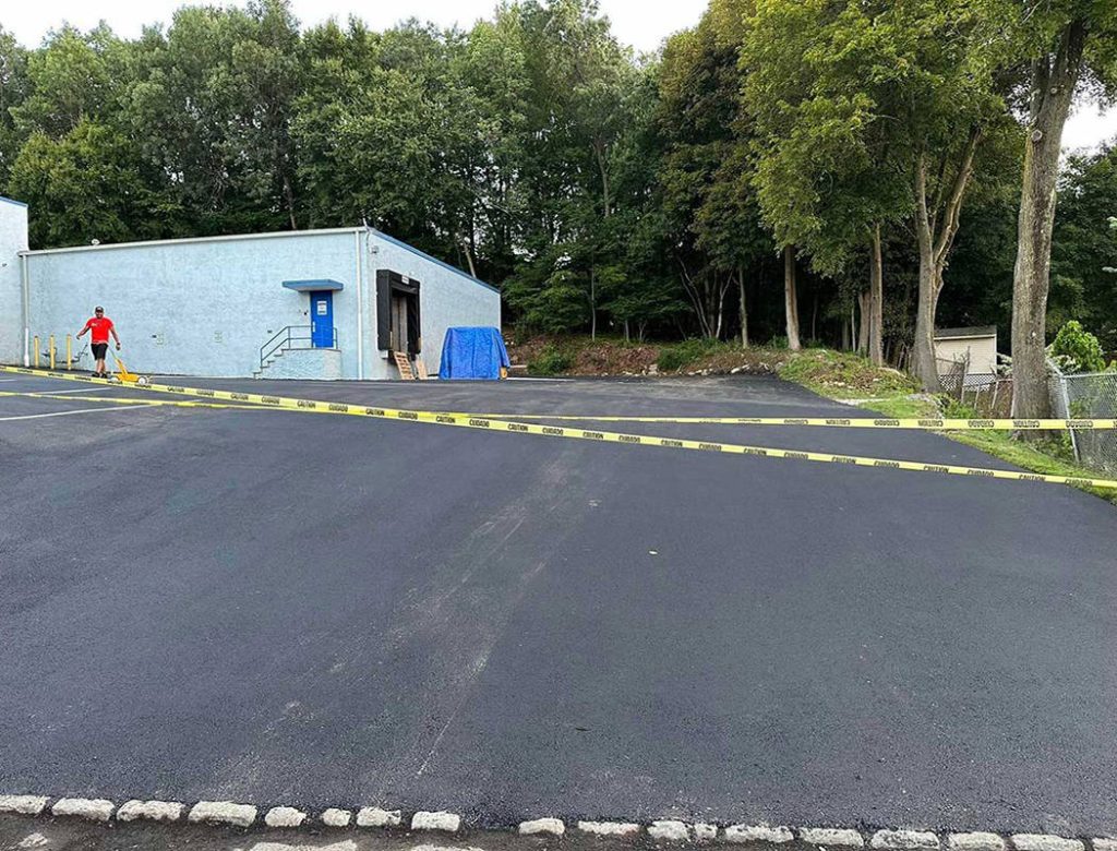 Freshly paved parking lot with caution tape, a person in red shirt, and trees in the background.