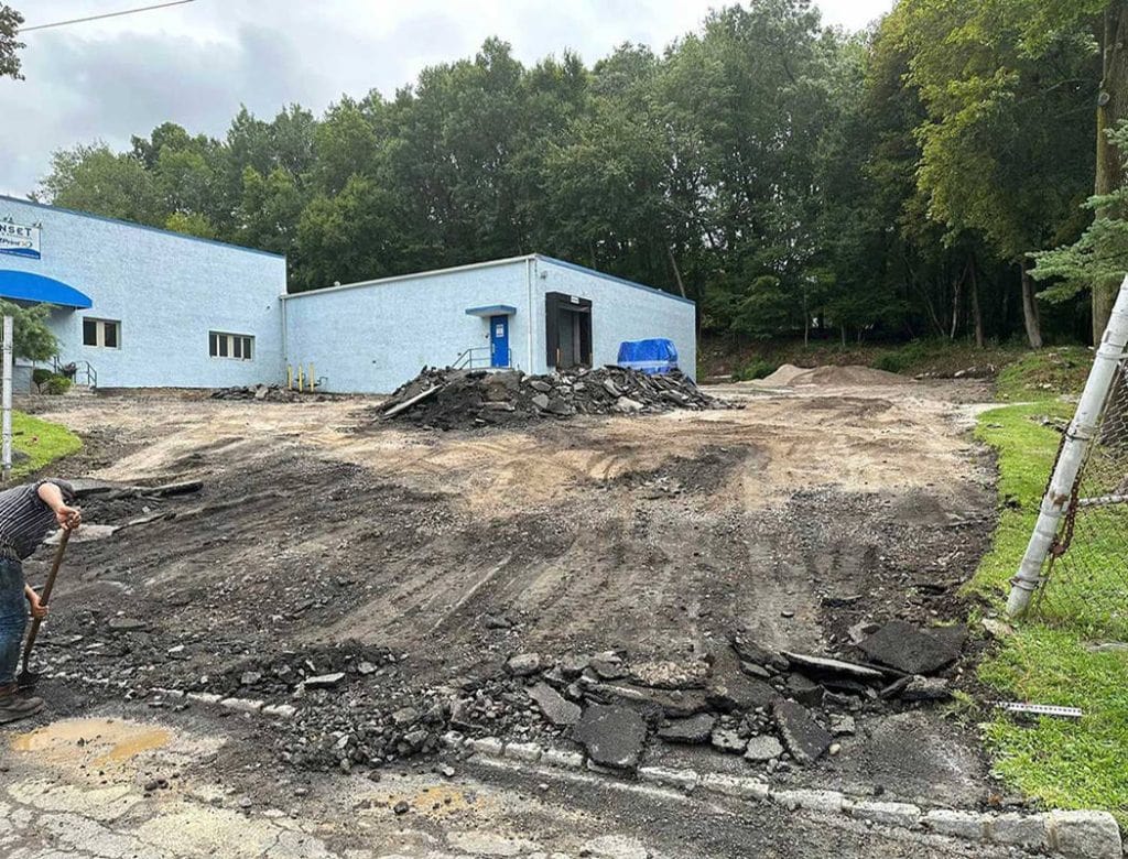 A worker shovels debris near a building with torn-up asphalt and dirt in front of it.