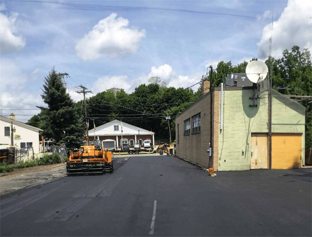 Freshly paved street with construction equipment, a beige building, and houses in the background under a blue sky.