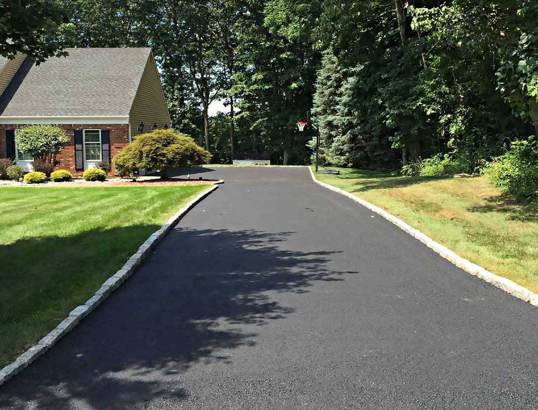 A newly paved black asphalt driveway curves past a brick house with green lawns and landscaping, bordered by stone edging and surrounded by tall trees.