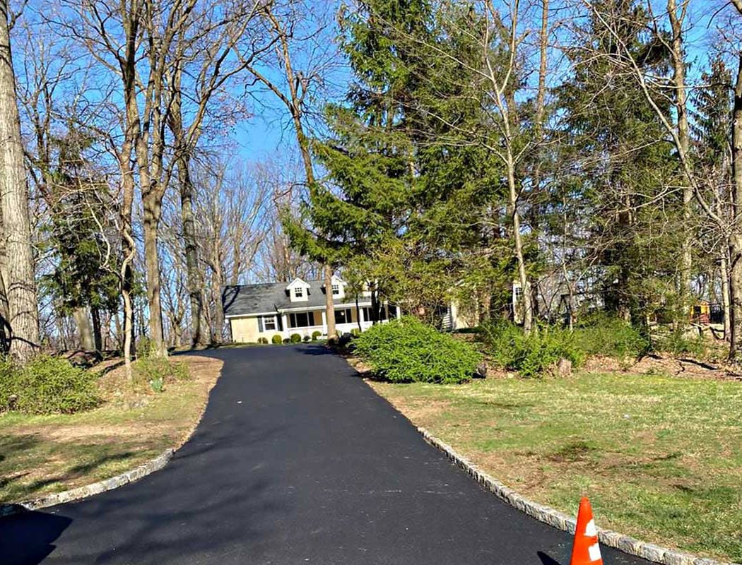 00006residential-1050x800 Residential paving: A paved driveway leads to a house with a front porch, surrounded by tall trees and green bushes; a single orange traffic cone is at the bottom right near the driveway.