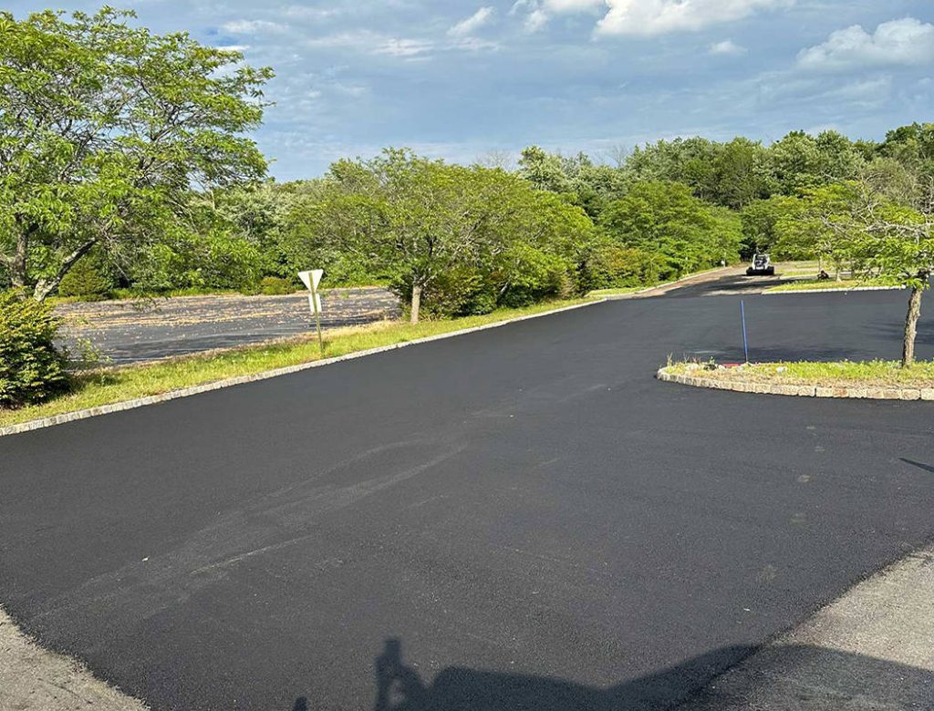 Freshly paved black asphalt road surrounded by green trees under a partly cloudy sky.