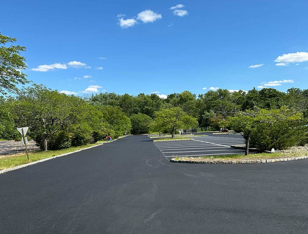 00009commercial-1050x800 A newly paved asphalt parking lot with a few marked parking spaces, surrounded by green trees under a blue sky with scattered clouds.