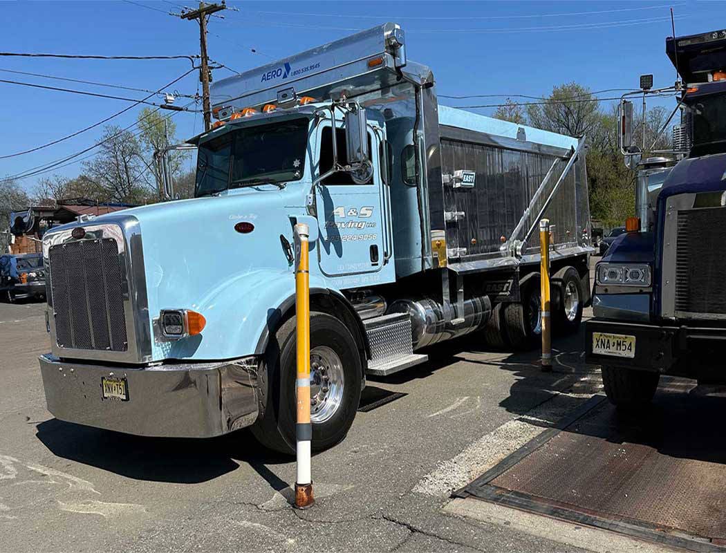 00009equipment-1050x800 A light blue dump truck is parked near a curb on a sunny day, surrounded by utility poles and other vehicles. Two yellow and white striped barriers stand in front of the truck.