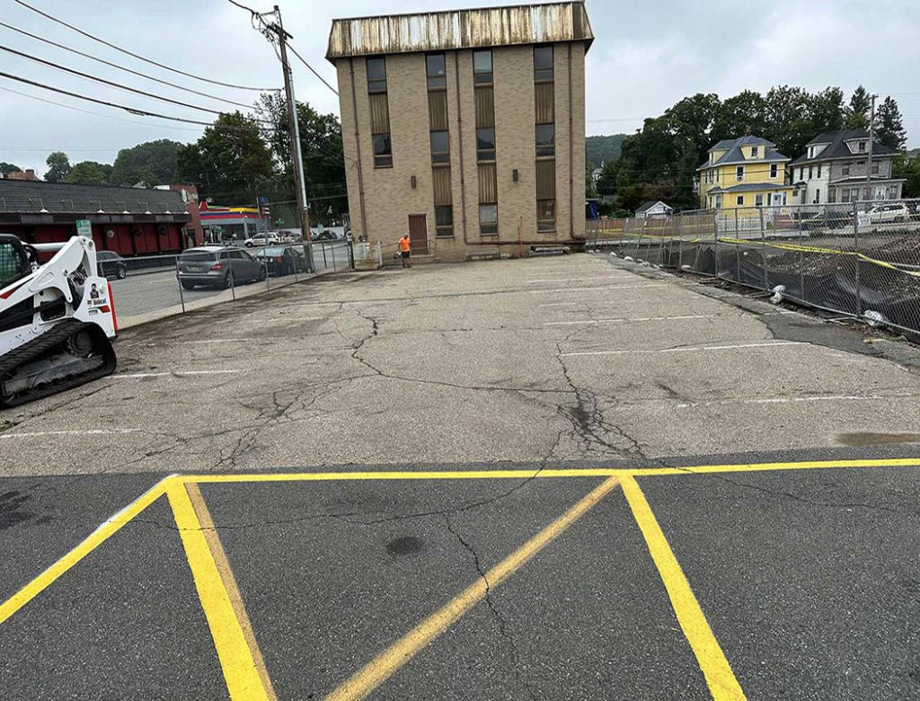 Empty cracked parking lot with faded yellow lines, bordered by fences, near a tall, worn building.