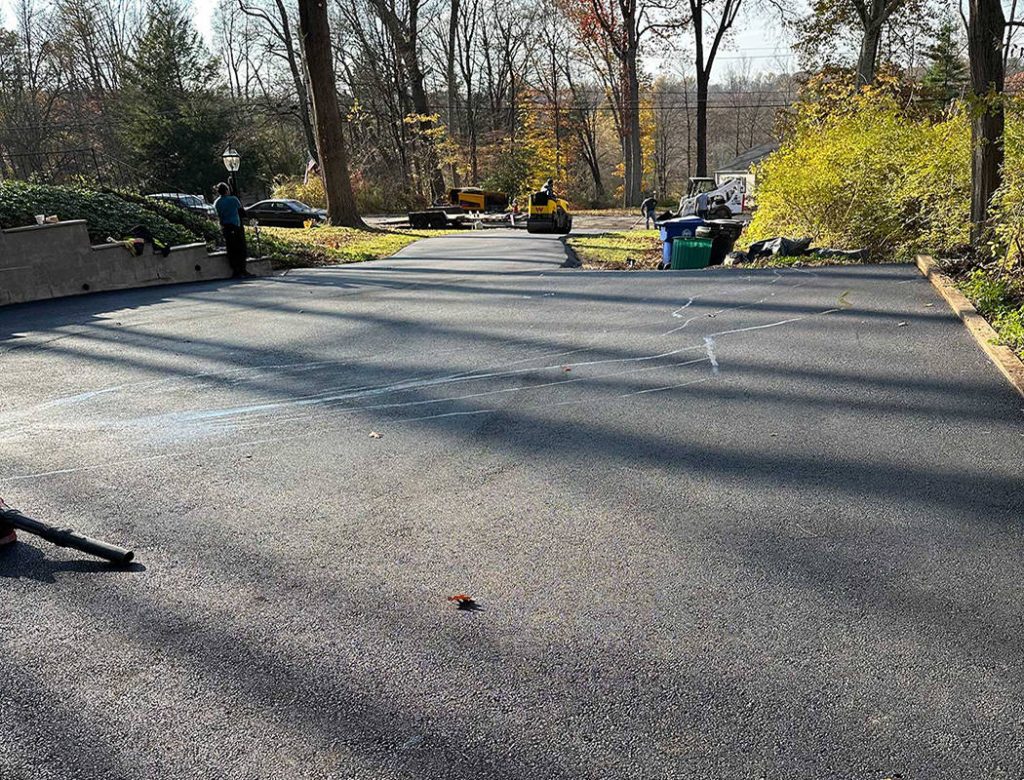 Freshly paved driveway with some chalk marks, surrounded by trees and parked vehicles in the background.
