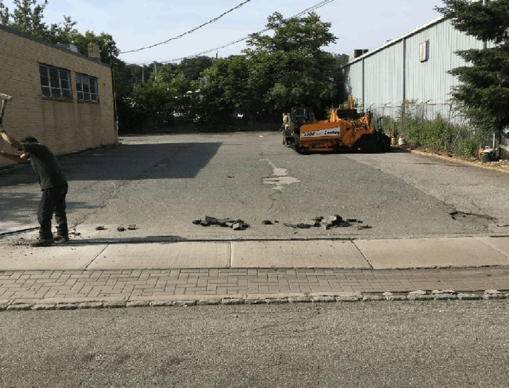 A worker breaks pavement near a parking lot; a yellow construction vehicle is in the background.