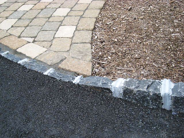 belgian-block-driveway A close-up of a border where asphalt meets a stone curb, with tan paving stones and wood mulch on the other side. The curb has white sealant in its joints.