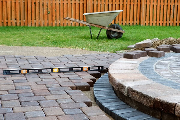 patio-construction A paved patio is being constructed with stone bricks. A level tool rests on the bricks, and a wheelbarrow is on the grass near a wooden fence in the background. Some stones are yet to be placed.
