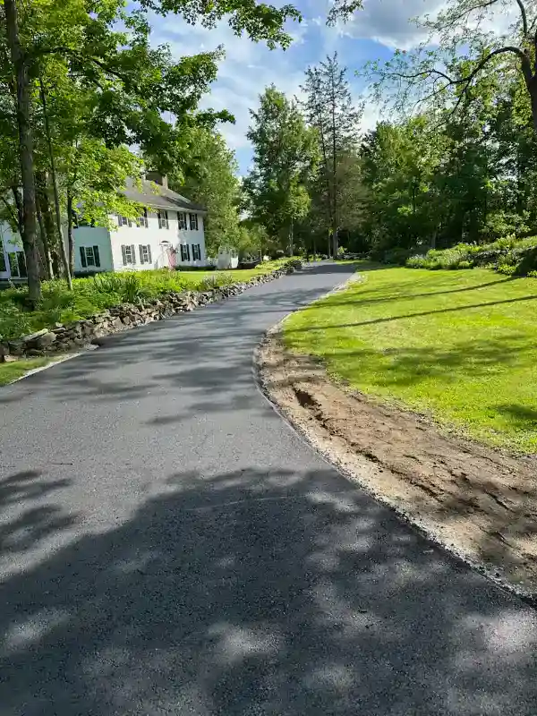 paving_morris-county_nj_cnfwjs A newly paved asphalt driveway curves through a green lawn toward a white house with black shutters, surrounded by trees and a rock-lined garden under a blue sky with scattered clouds.