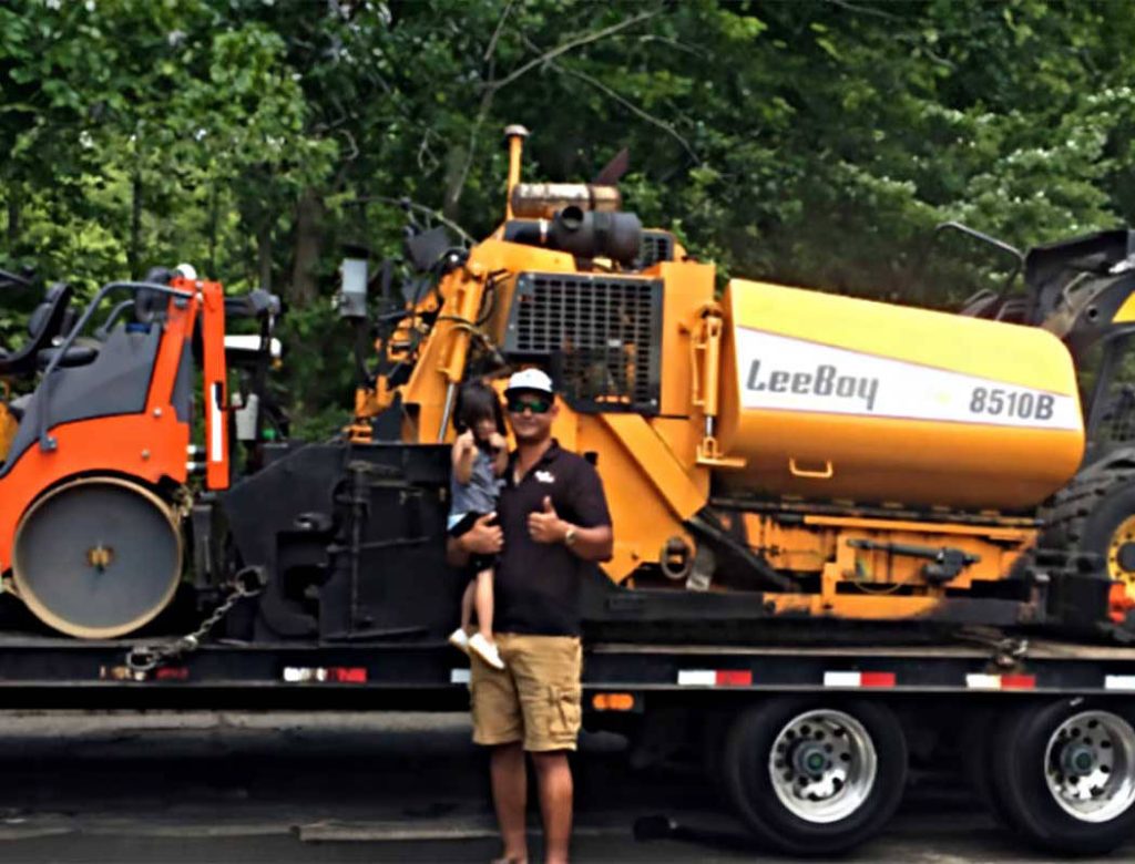 Man holding a child stands in front of a yellow LeeBoy 8510B paver and an orange roller on a trailer.