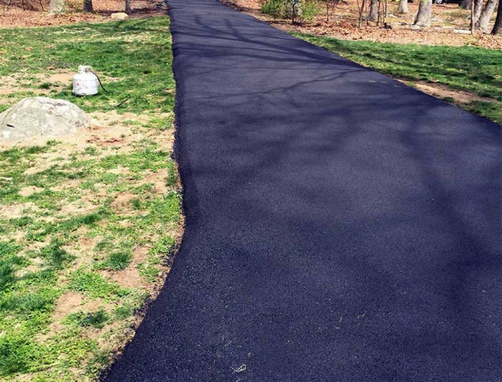Freshly paved black asphalt path curving through a grassy area with trees and a large rock nearby.