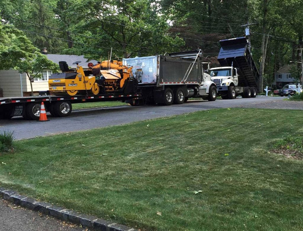 Two dump trucks and construction equipment parked on a residential street near a grassy yard.