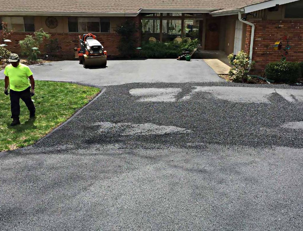 Worker paving a driveway with fresh asphalt near a brick house, while another operates a machine.