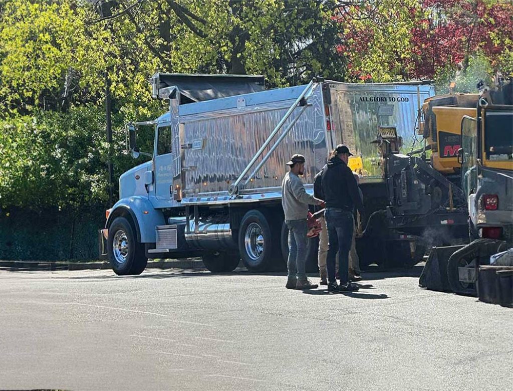 Three workers stand near a shiny silver dump truck and construction equipment on a sunny day.
