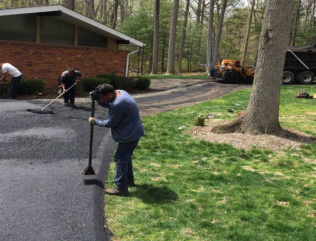 Workers paving a driveway with fresh asphalt near a brick house surrounded by trees and grass.