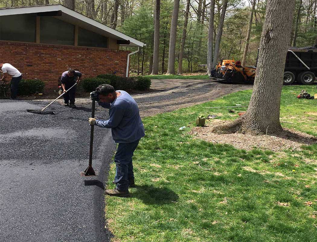 00003residential-1050x800-1 Three workers are paving a driveway with fresh asphalt outside a brick house surrounded by trees and grass. One man smooths the edge, while two others work further back with tools and a paving machine.