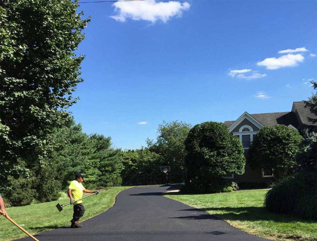 A worker in a yellow shirt paves a driveway near a suburban house on a sunny day.