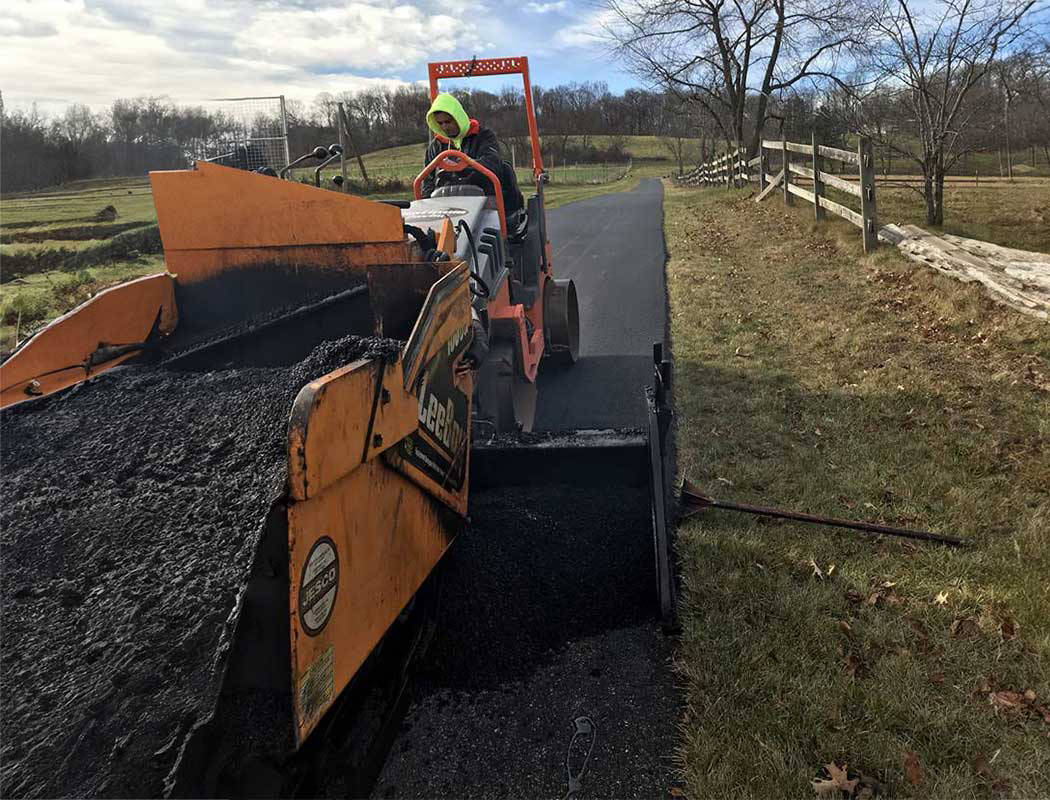 00004equipment-1050x800-1 A worker in a neon safety hoodie operates an asphalt paving machine, laying fresh blacktop on a rural road bordered by grass, wooden fences, and leafless trees under a partly cloudy sky.