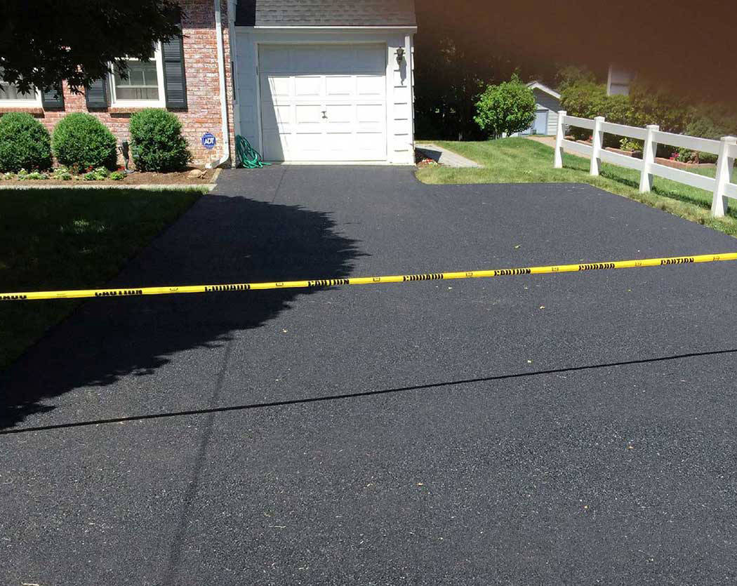 00004residential-1050x835-1 A freshly paved driveway in front of a house is blocked off with yellow caution tape, preventing entry. There's a white garage door, green bushes, and a white fence alongside the driveway.