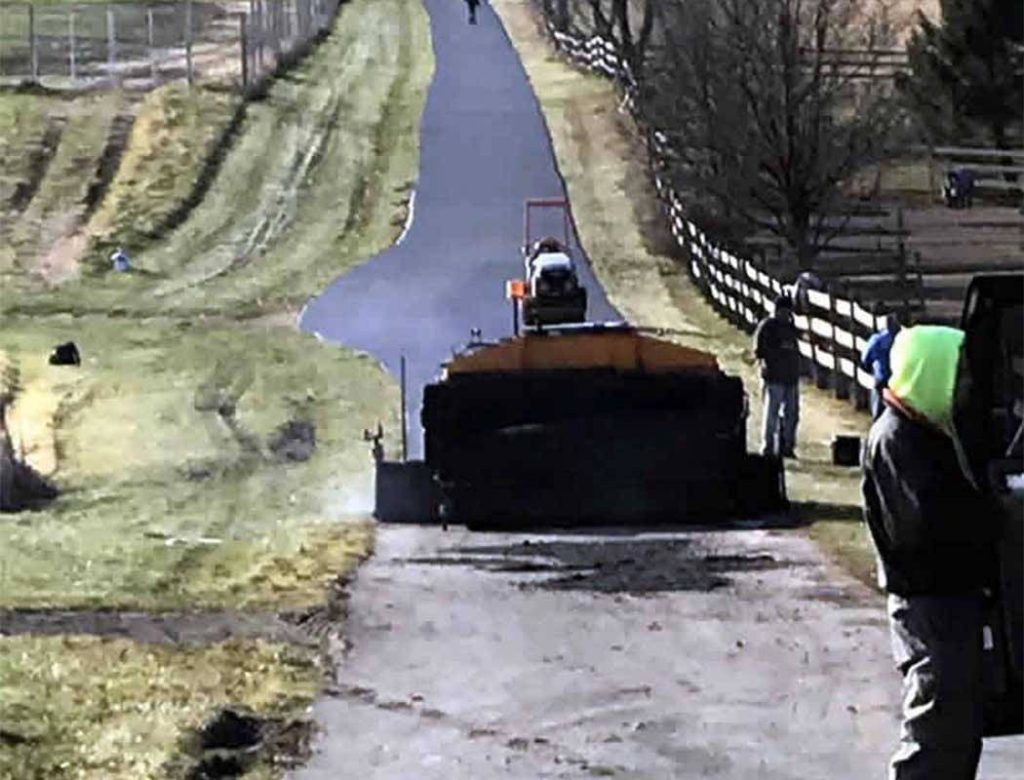 A machine paves a narrow road as workers stand nearby on a rural, fenced-in path.