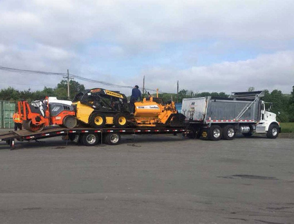 A flatbed truck is hauling three pieces of yellow construction equipment on a trailer in a parking lot.