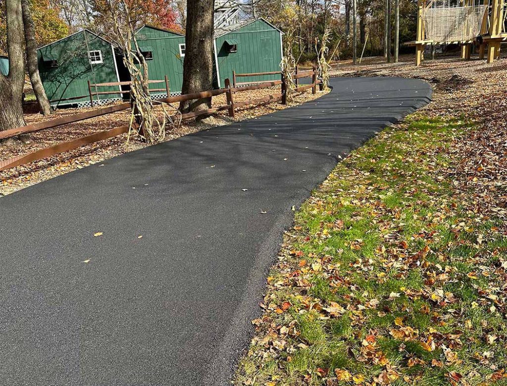 A newly paved asphalt path curves past green sheds, trees, and fallen autumn leaves.