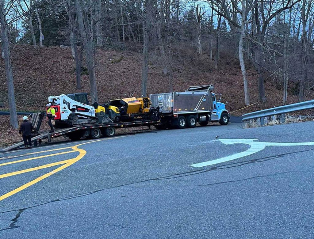 A truck with construction equipment on a trailer is parked on a road near a curve, with two people nearby.