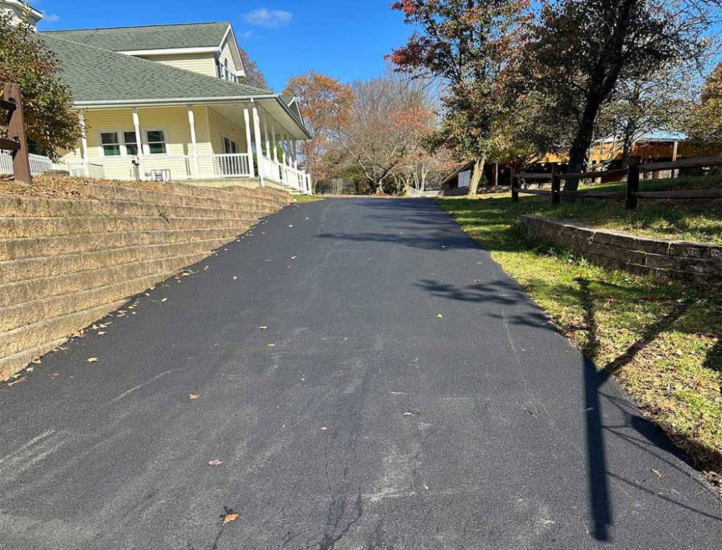 Paved driveway leading up to a house with a porch, bordered by stone walls and trees under a blue sky.