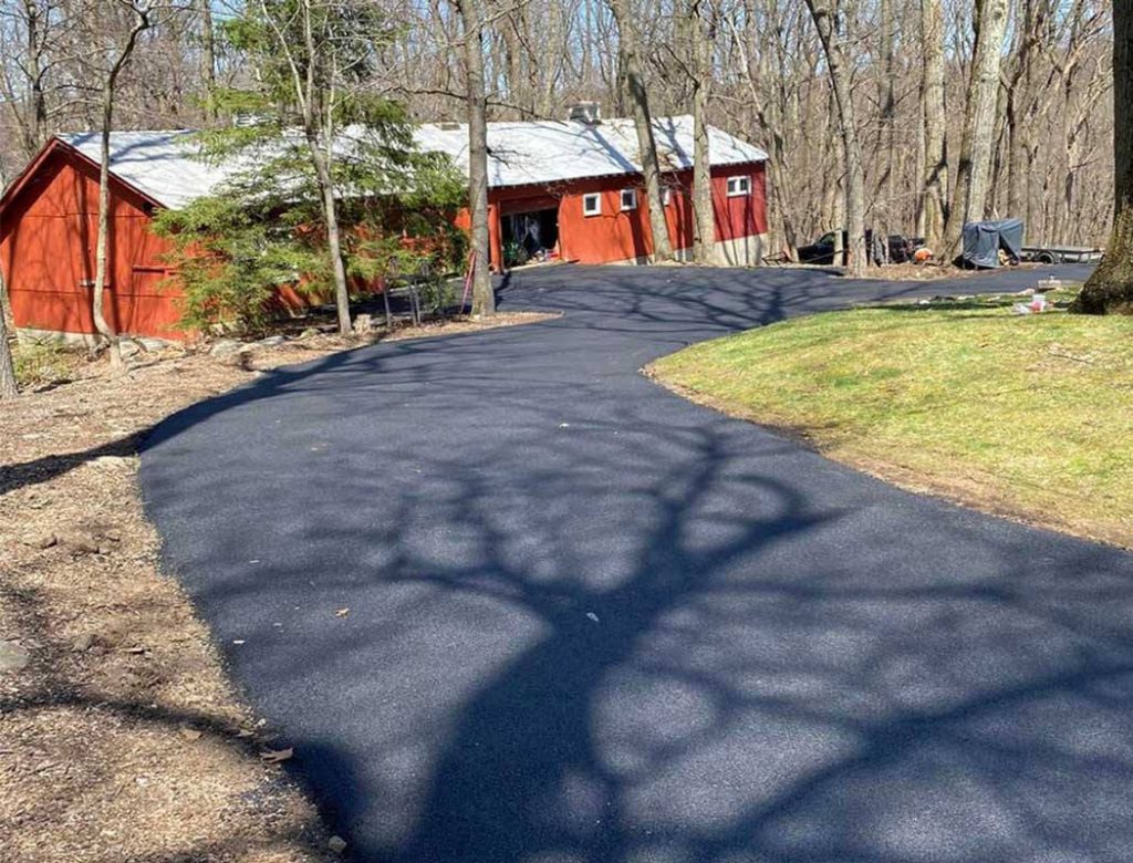 A newly paved driveway leads to a red house surrounded by leafless trees and patches of grass.