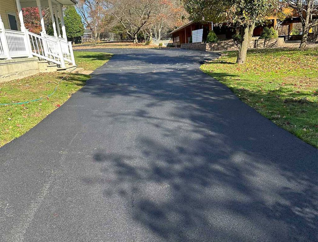 Freshly paved asphalt driveway curves past a house, with grass and trees on either side under clear skies.