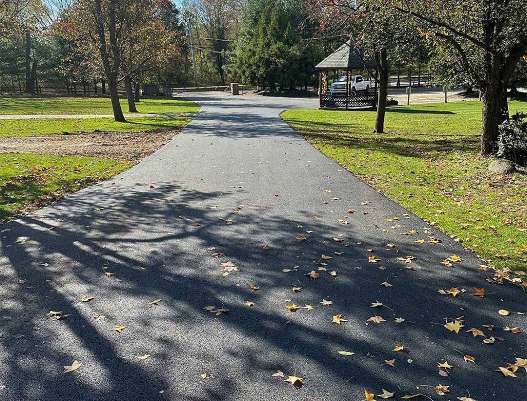 A paved path with scattered autumn leaves and trees leading to a gazebo in a sunny park.