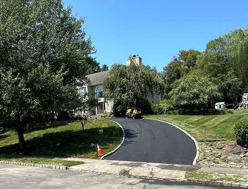 A freshly paved black asphalt driveway curves up to a house surrounded by green trees and grass. An orange traffic cone sits at the driveway entrance, and a small yellow roller is parked halfway up the drive.