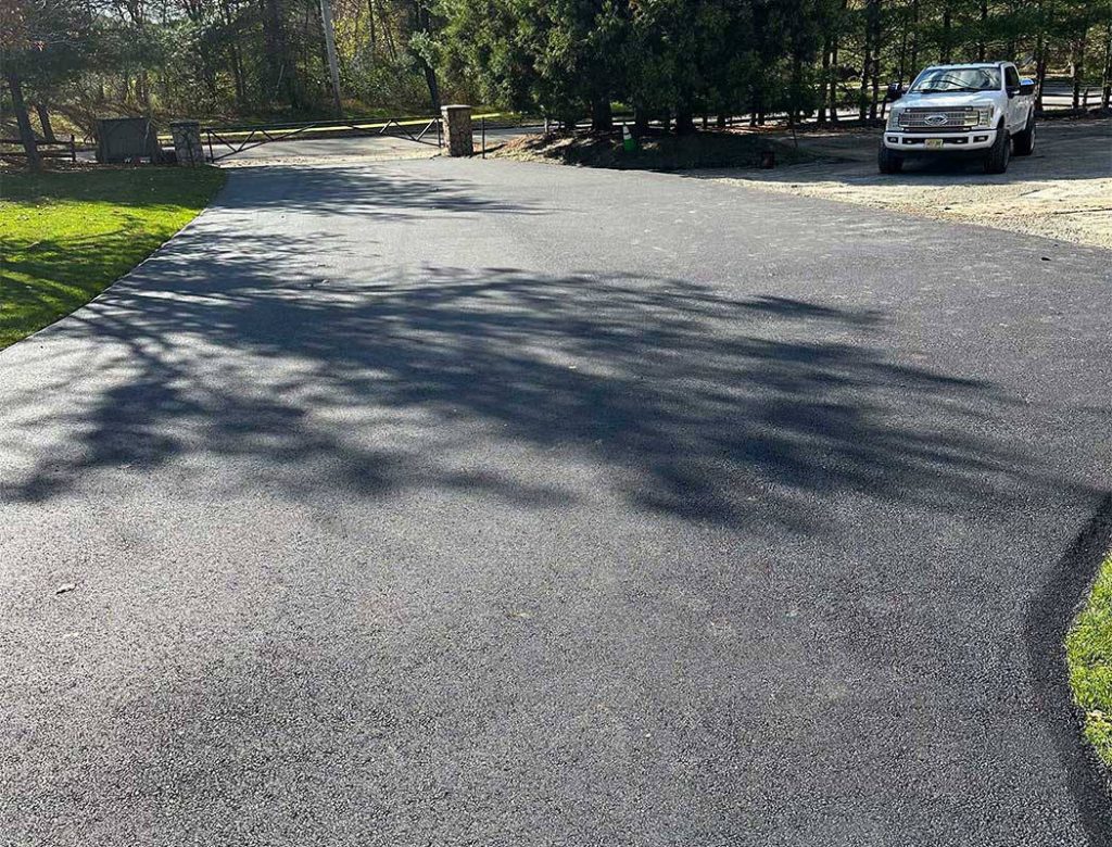 Freshly paved asphalt driveway with a white pickup truck parked nearby and trees casting shadows.