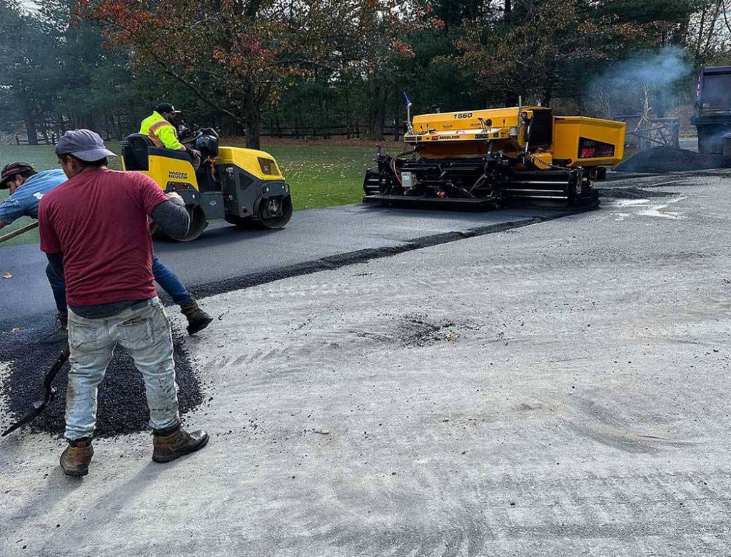 Workers paving a road with asphalt; a yellow paver and roller are being used on a partially finished surface.