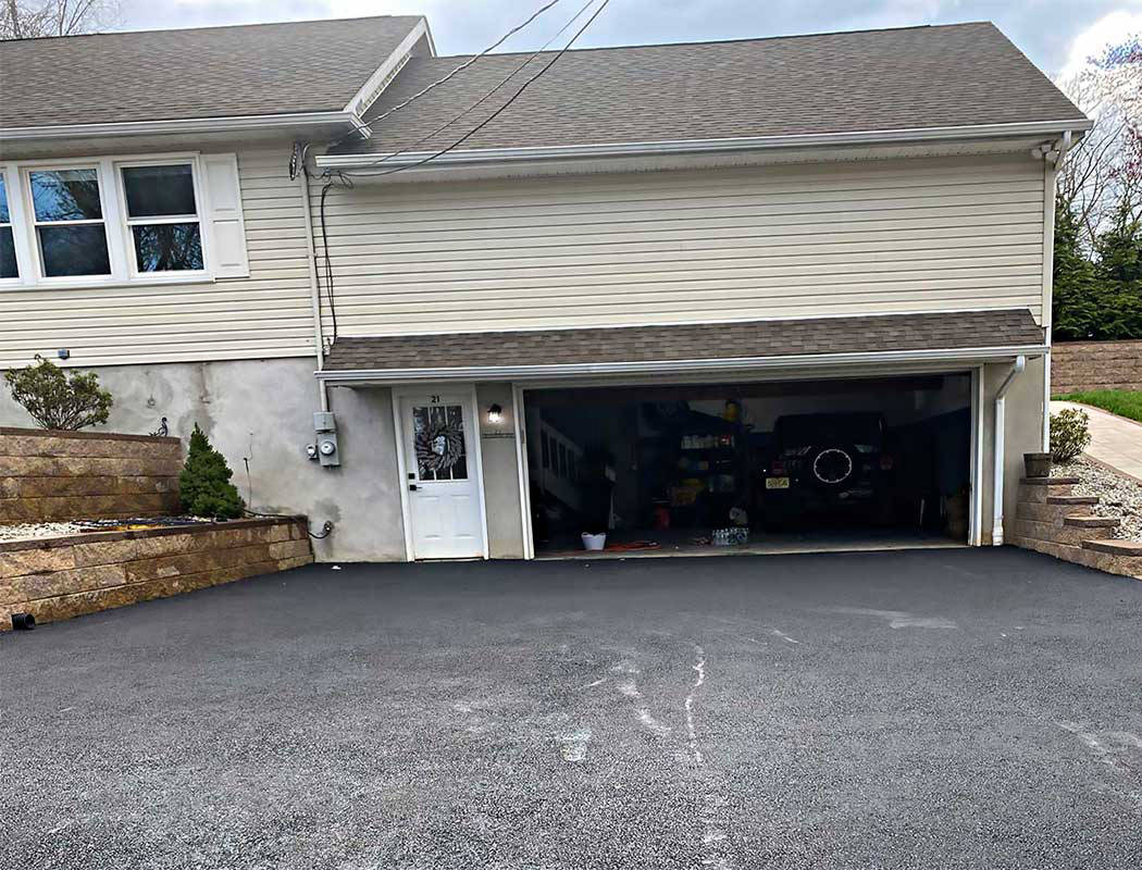 A house with a double garage door open, showing a black vehicle and various items inside. The driveway is freshly paved with dark asphalt and has stone retaining walls on both sides.