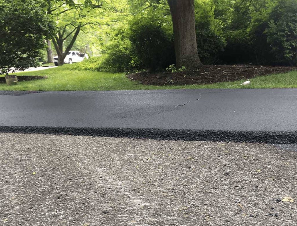 Freshly paved asphalt driveway with a distinct edge meets an older gravel surface, surrounded by green trees.