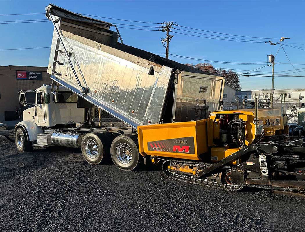 A dump truck unloads asphalt into a yellow road paving machine at a construction site on a sunny day, with buildings and power lines in the background.