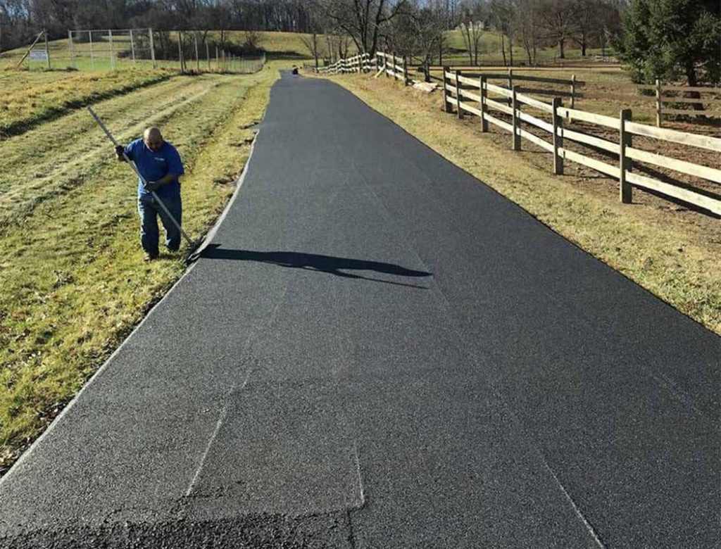 Person smoothing freshly laid asphalt on a rural driveway beside a wooden fence and grassy field.