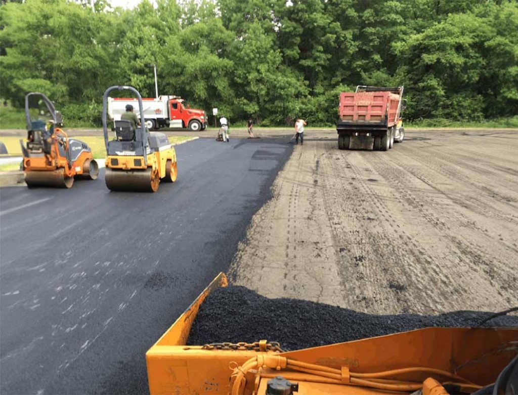 Workers and construction vehicles paving a road with fresh asphalt near a green, wooded area.