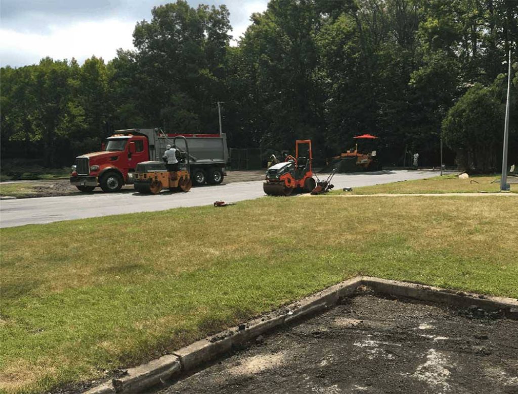 Two workers use paving equipment near a dump truck on a road beside a grassy area with trees in the background.
