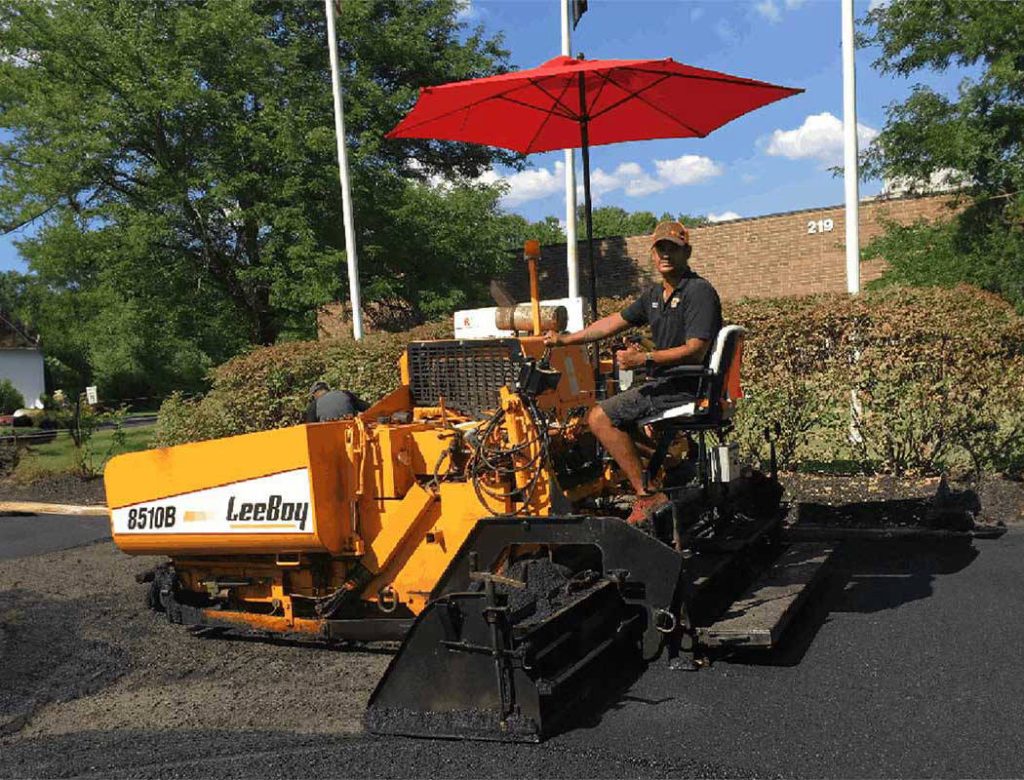 A man operates an orange paving machine with a red umbrella shade on a sunny day.