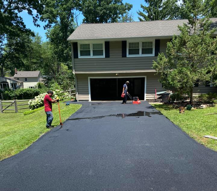 Two people are resurfacing a driveway in front of a gray two-story house; one is spreading material with a long tool while the other works near the garage on a sunny day.