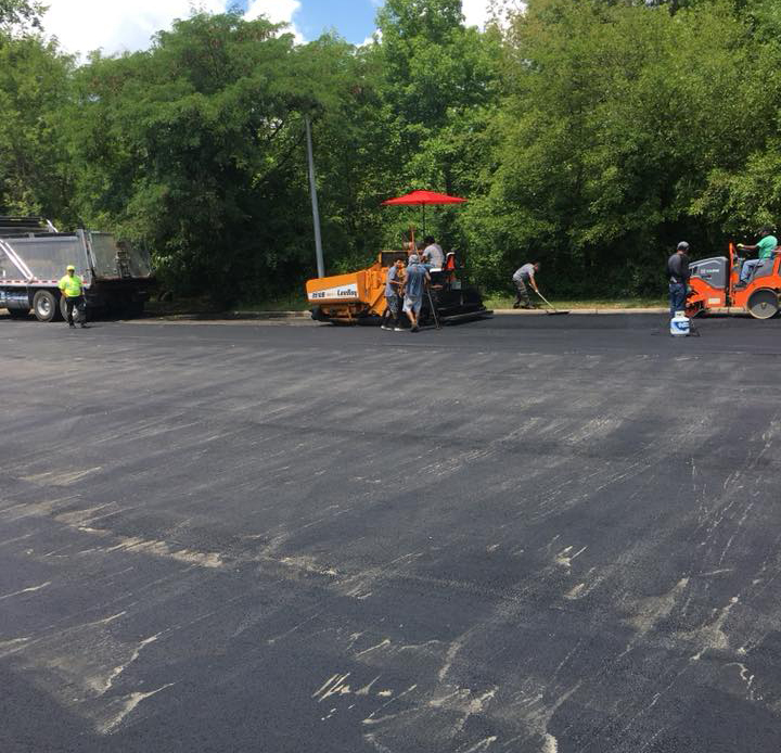 167403275_1353938718307433_7313799505238190199_n Workers are paving a parking lot with asphalt, using construction equipment including a paver and roller, near a dump truck. Trees and greenery line the background.