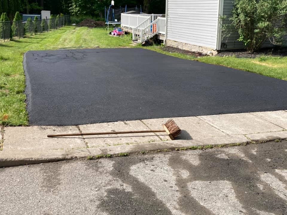 A freshly paved black asphalt driveway leads to a house with a porch. A broom lies across the concrete sidewalk at the edge of the driveway. Grass and a fence border the area.