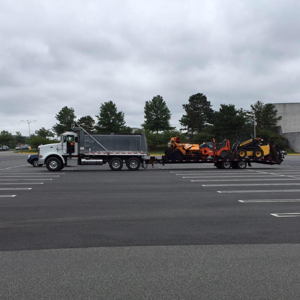 493817686_2461442217557072_3655991482271996455_n A large white dump truck is parked in an empty lot, towing a trailer loaded with two orange and black construction vehicles under a cloudy sky. Trees and a gray building are visible in the background.